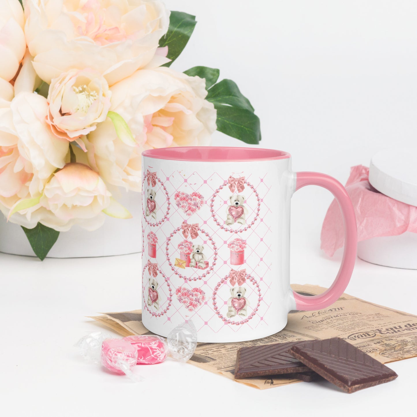 White mug with pink interior and handle, featuring cute illustrations, on a white surface with flowers and chocolate.