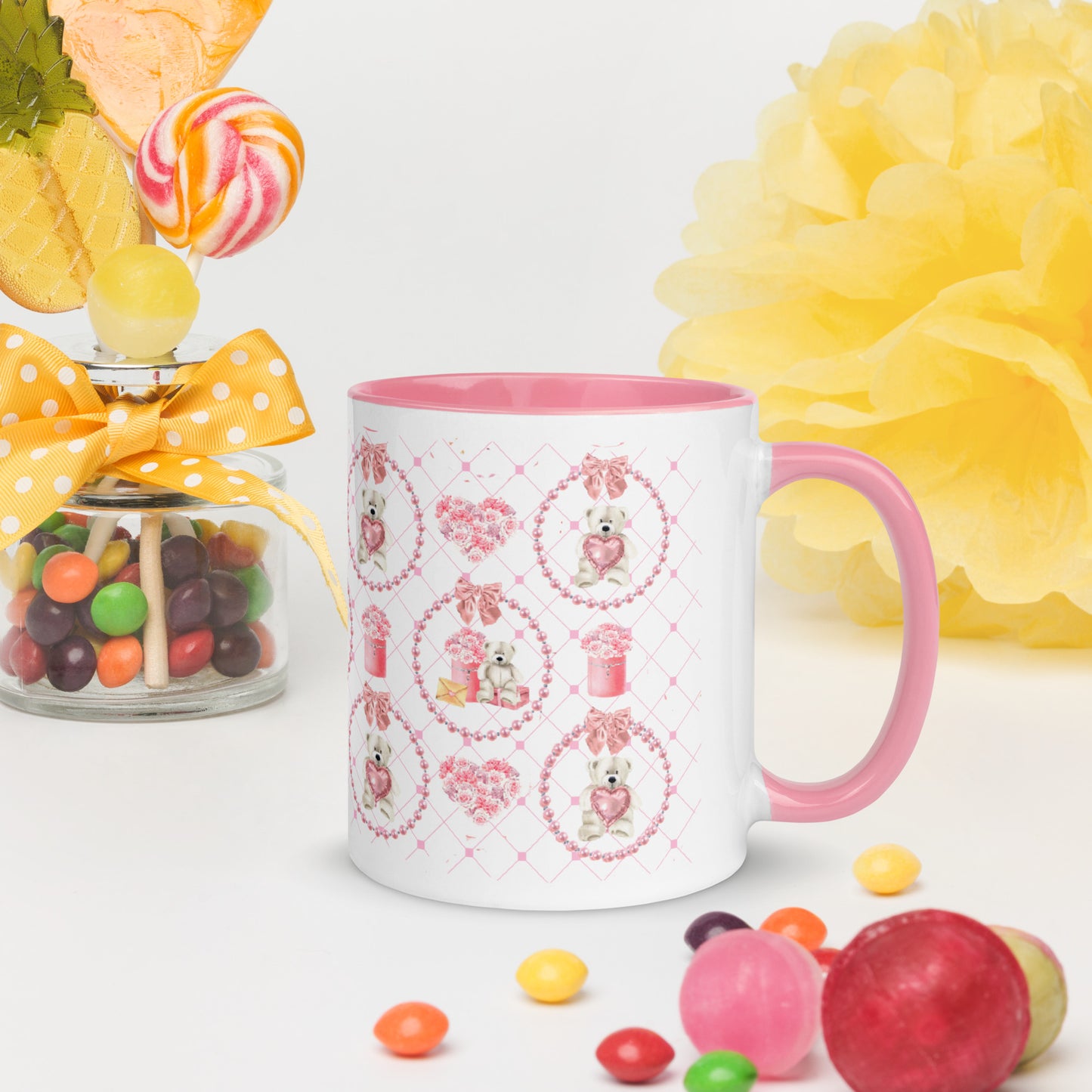White mug with pink interior and handle, featuring teddy bear designs, surrounded by colorful candies and flowers.