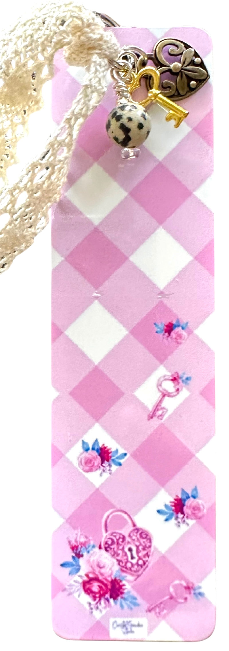 A lightweight aluminum bookmark with vintage lace, charms, and a Jasper bead, displayed against a pink checkered background.